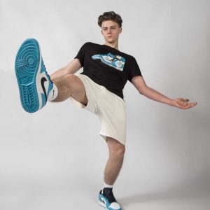 Featured image of a young guy kicking his foot out, wearing the Baller Boys Dunk Tee t-shirt and Baseline shorts in Ecru